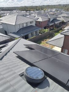A rooftop view of suburban houses with tiled roofs, featuring solar panels and a round Solatube solar roof ventilator in the foreground.